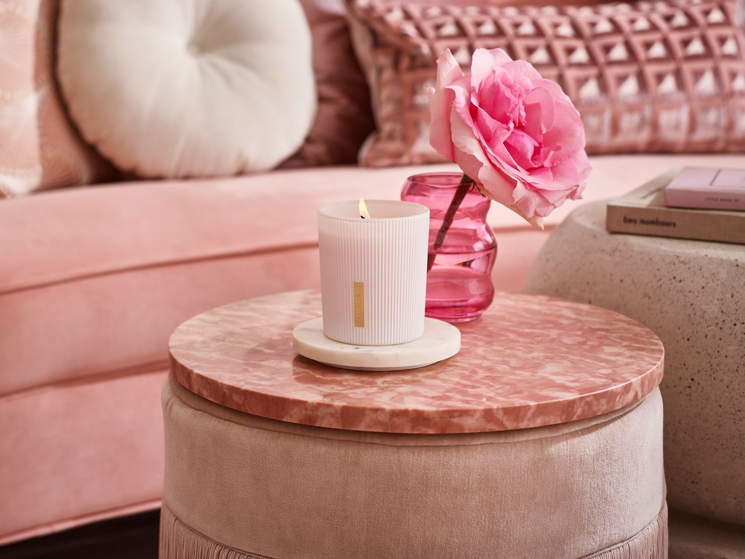 Na okrągłym stoliku w odcieniach różu stoi biała świeca, różowy wazon z różą i książki, tworząc przytulną atmosferę.