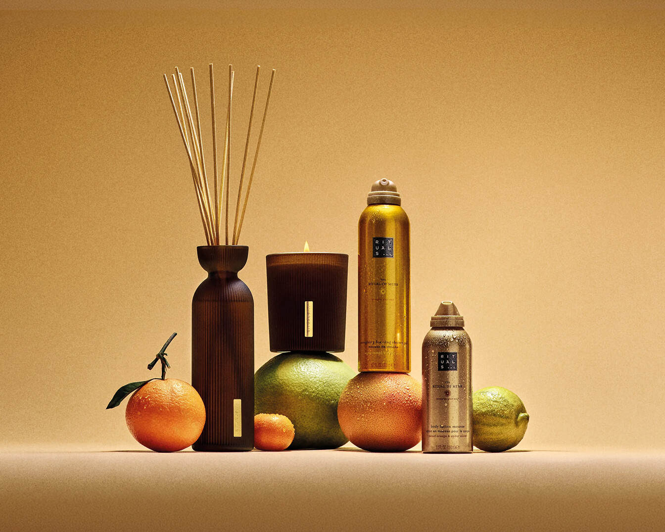 Na beżowym tle znajdują się produkty Rituals, takie jak świeca, dyfuzor zapachowy, złote i brązowe butelki oraz owoce cytrusowe.