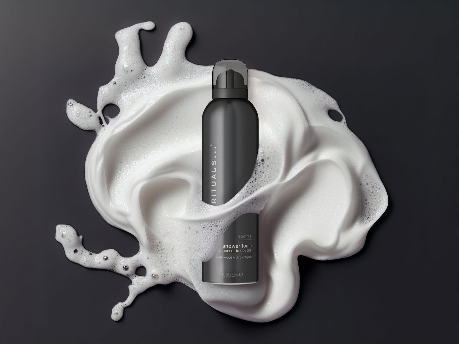 Czarna butelka pianki pod prysznic Rituals Homme otoczona białą pianą na ciemnym tle.