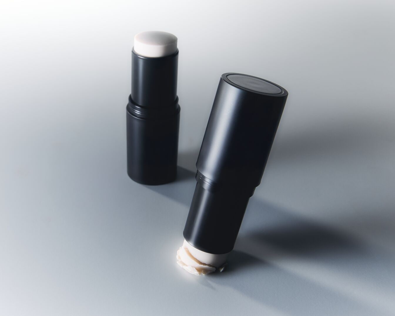 Ecco due stick di fondotinta in confezione nera opaca, uno in piedi e l'altro appoggiato di lato con un po' di prodotto color crema chiaro fuoriuscito.