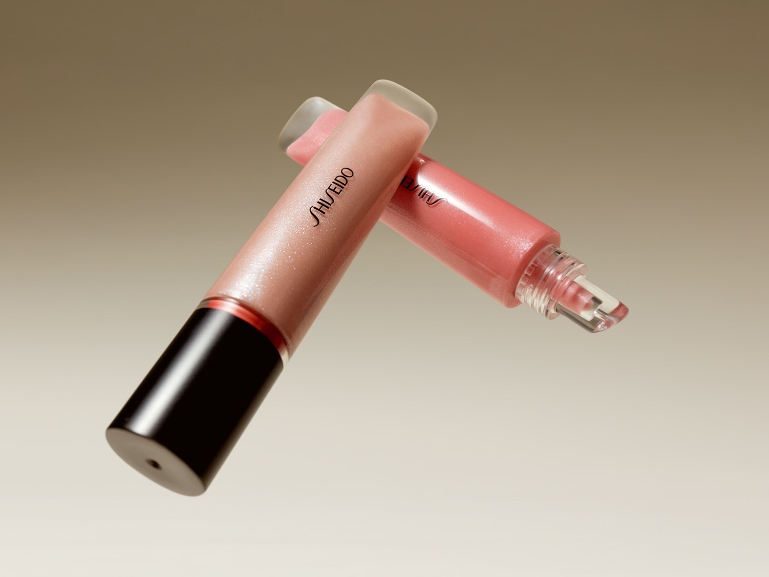 Két Shiseido szájfény fekszik egymáson; az egyik bézs, a másik rózsaszín, mindkettő fekete kupakkal és átlátszó applikátorral.