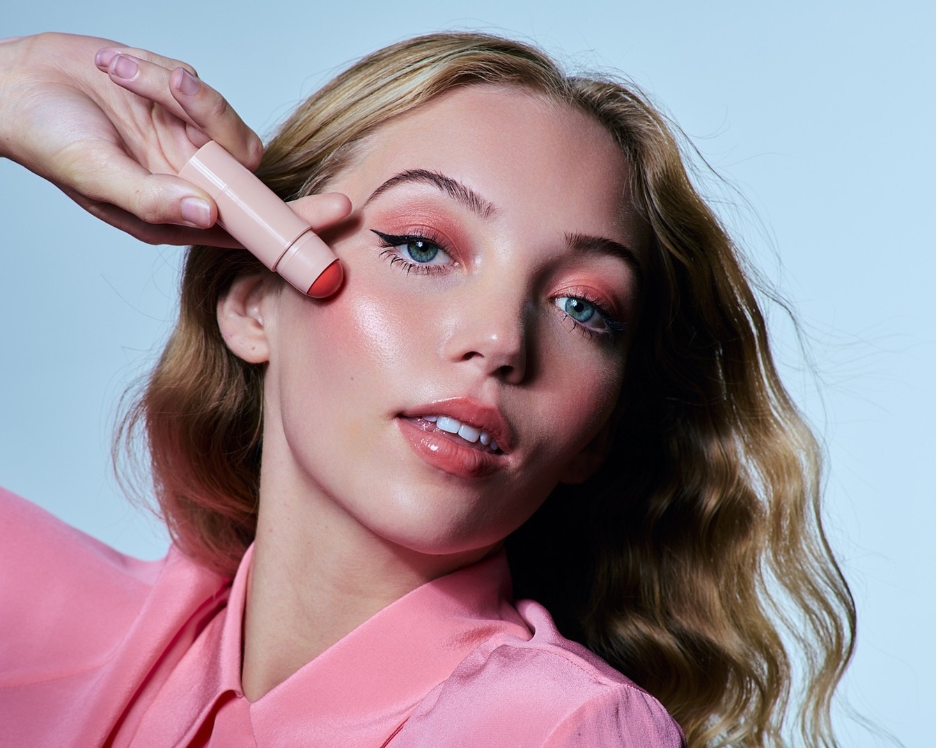 Egy rózsaszín blúzt viselő nő egy rózsaszín krémes pirosítót tart az arcához, kiemelve a rózsaszín szemhéjfestékkel és fényes ajkakkal kiegészített ragyogó sminkjét.
