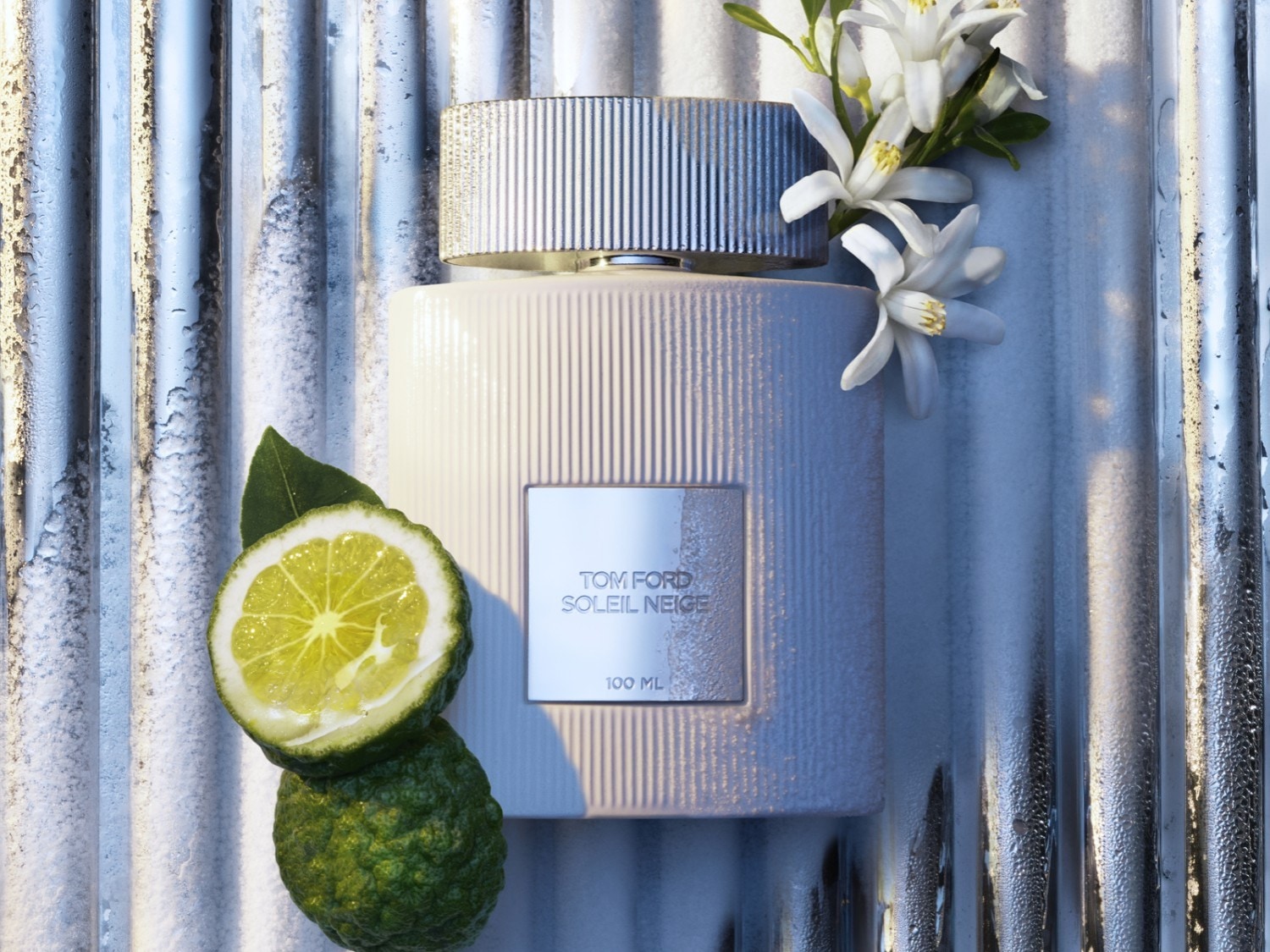 Biały flakon perfum Tom Ford Soleil Neige o pojemności 100 ml, ozdobiony kwiatami i limonką, na tle srebrnych rurek.