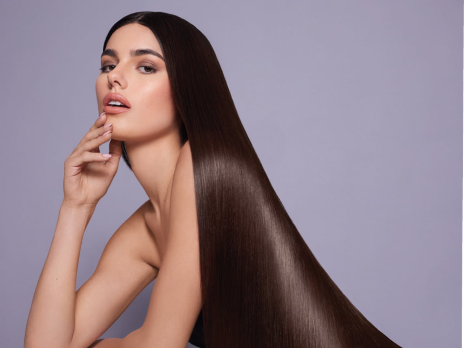 Eine Frau mit langen, glänzenden, dunkelbraunen Haaren posiert vor einem violetten Hintergrund und berührt sanft ihr Gesicht.