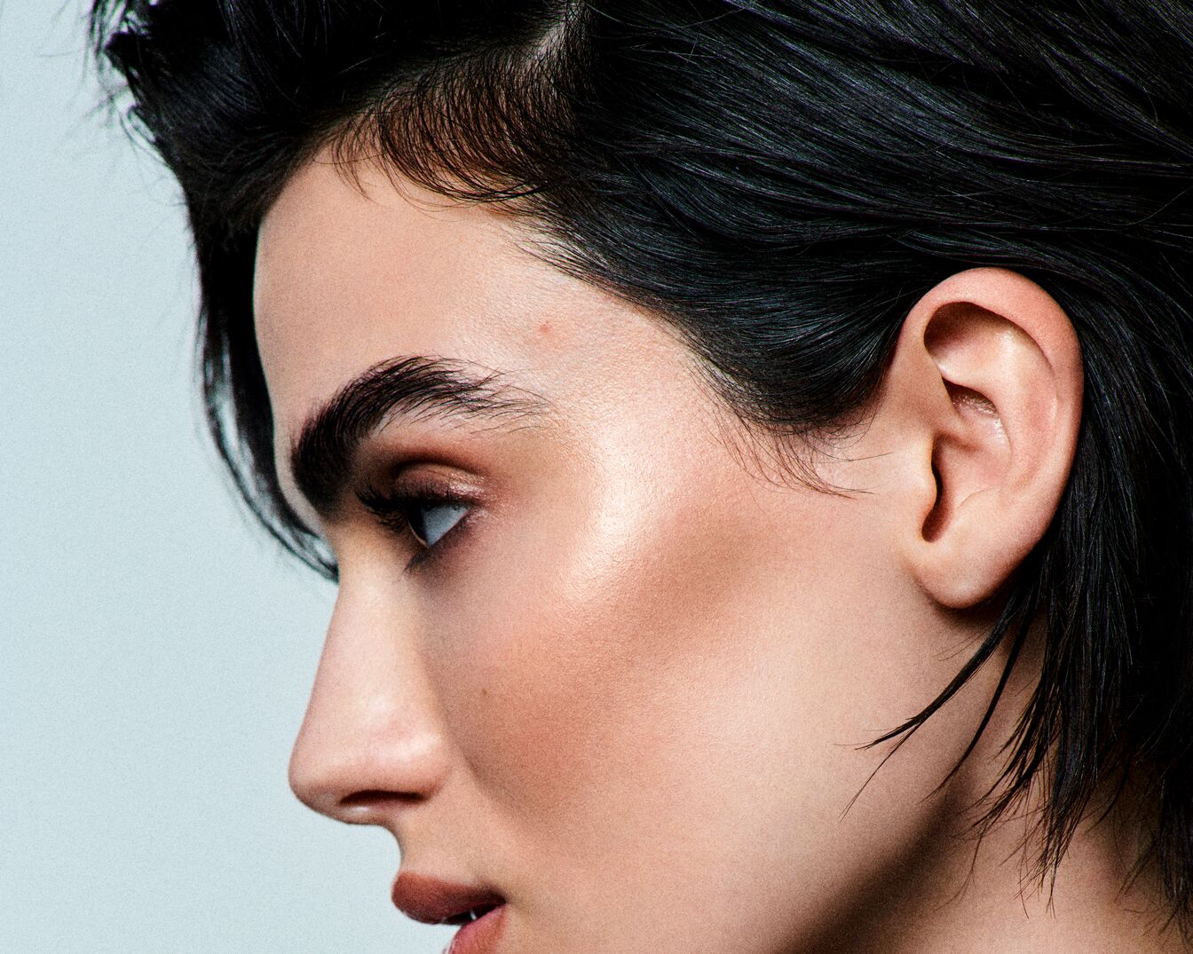 Primo piano di un viso di profilo con pelle luminosa, sopracciglia folte e capelli neri lucidi, che mette in risalto i dettagli del trucco e la texture della pelle.