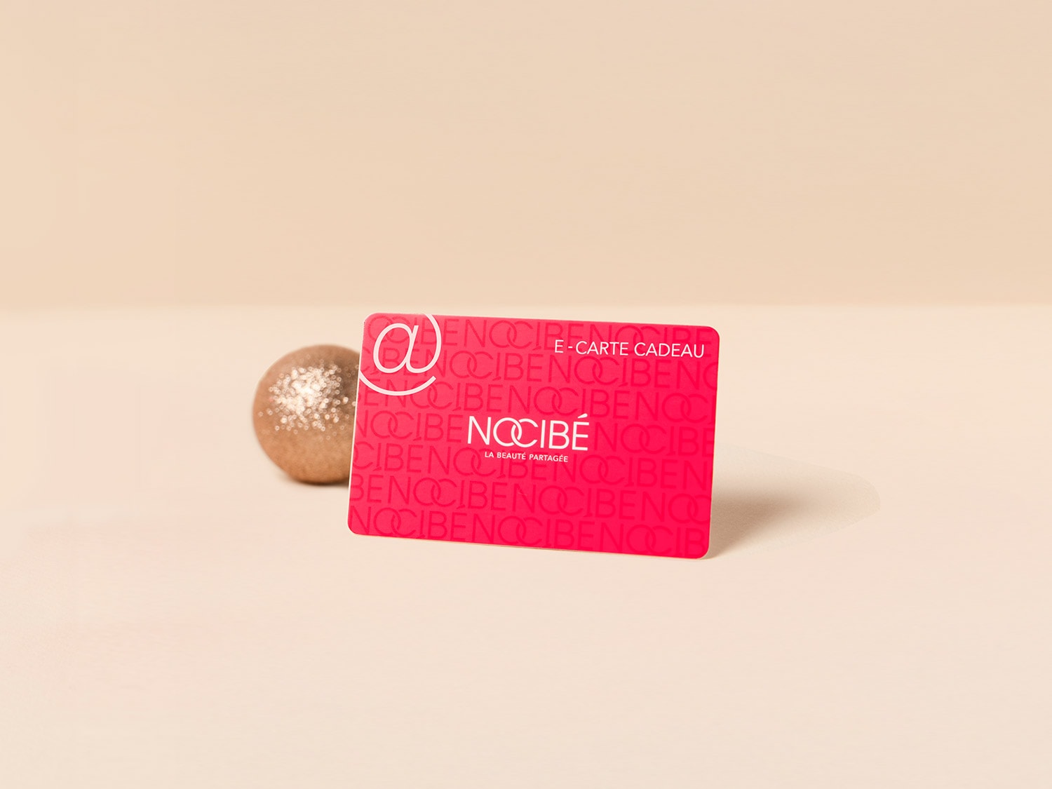 Voici une carte cadeau Nocibé rose avec le logo Nocibé répété et une boule décorative dorée à côté.