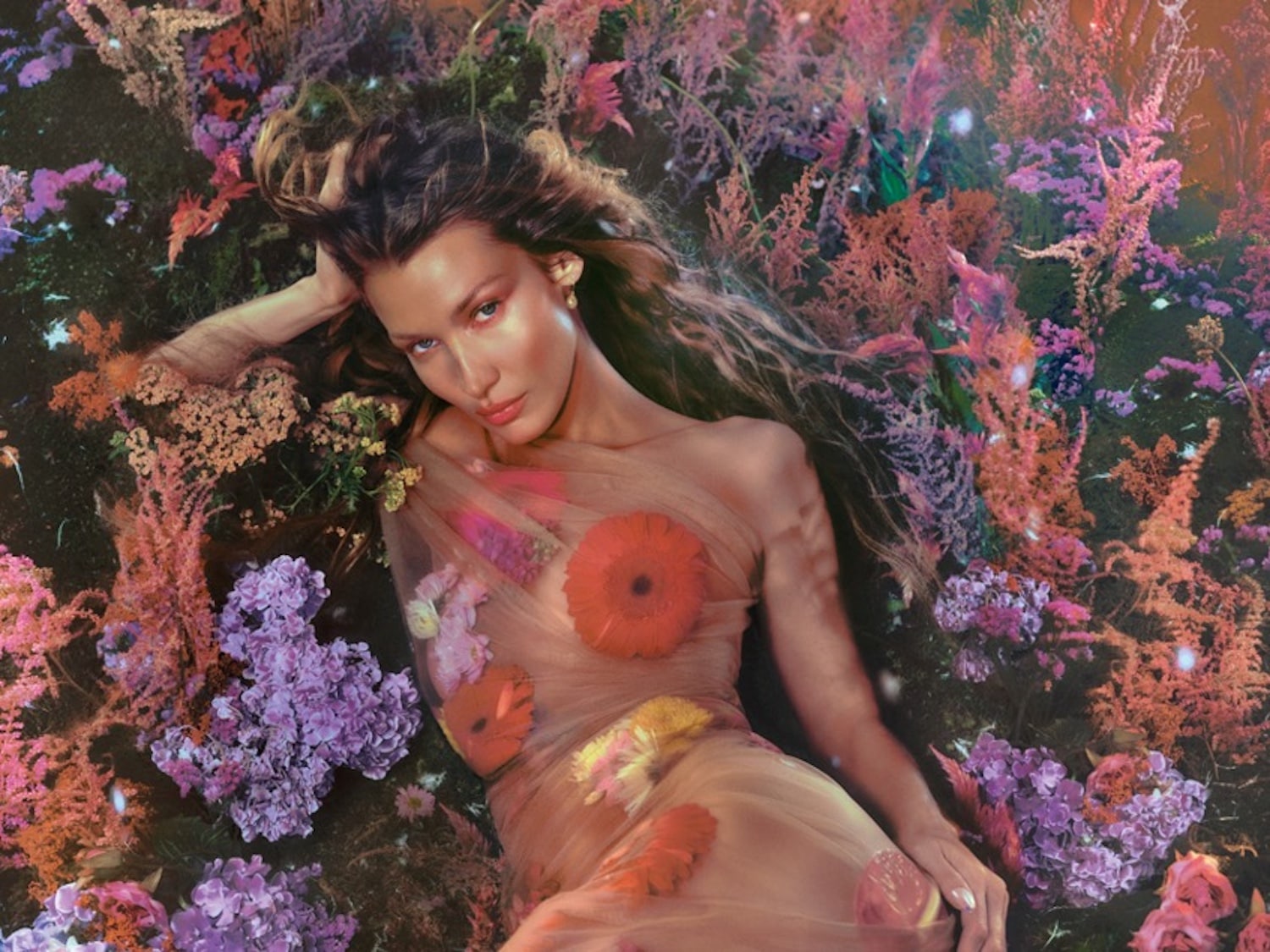 Nuotraukoje moteris guli apsupta spalvingų gėlių, vilkėdama permatomą suknelę su gėlių aplikacijomis, sukuriančią gamtos ir grožio harmoniją.