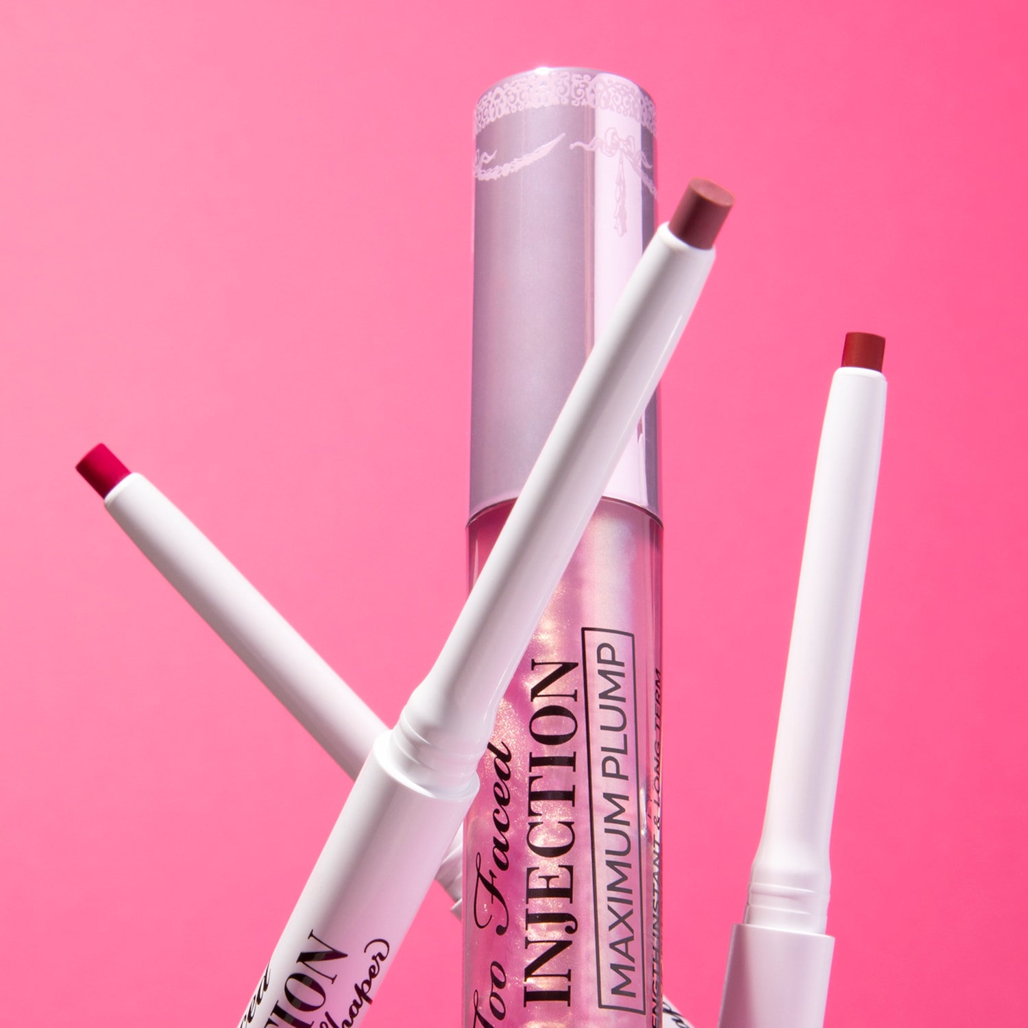 Vor einem pinkfarbenen Hintergrund steht ein Lip Injection Maximum Plump Lipgloss umgeben von drei Liplinern.