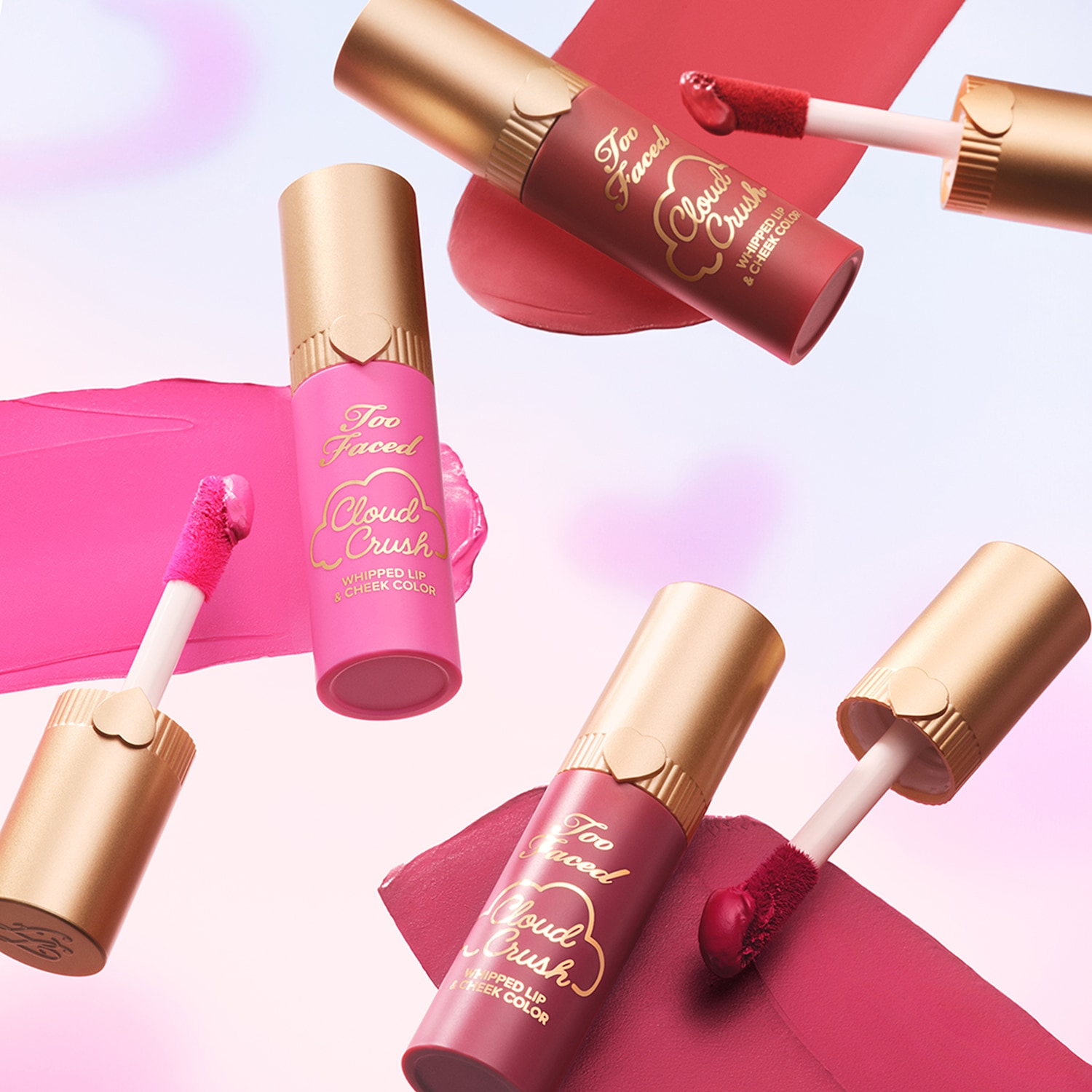 Mehrere Too Faced Cloud Crush Whipped Lip & Cheek Color Blushes in verschiedenen Pink- und Rottönen mit goldenen Deckeln vor einem wolkenähnlichen Hintergrund.