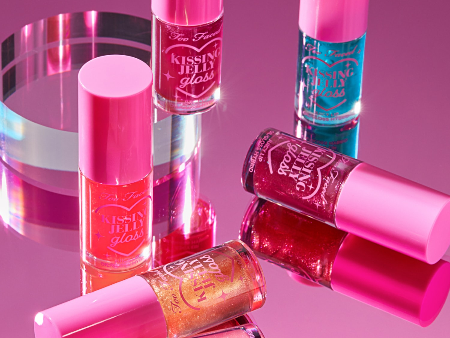 Verschiedene Farbtöne von Too Faced Kissing Jelly Gloss mit rosa Deckeln, die auf einer reflektierenden rosa Oberfläche präsentiert werden.