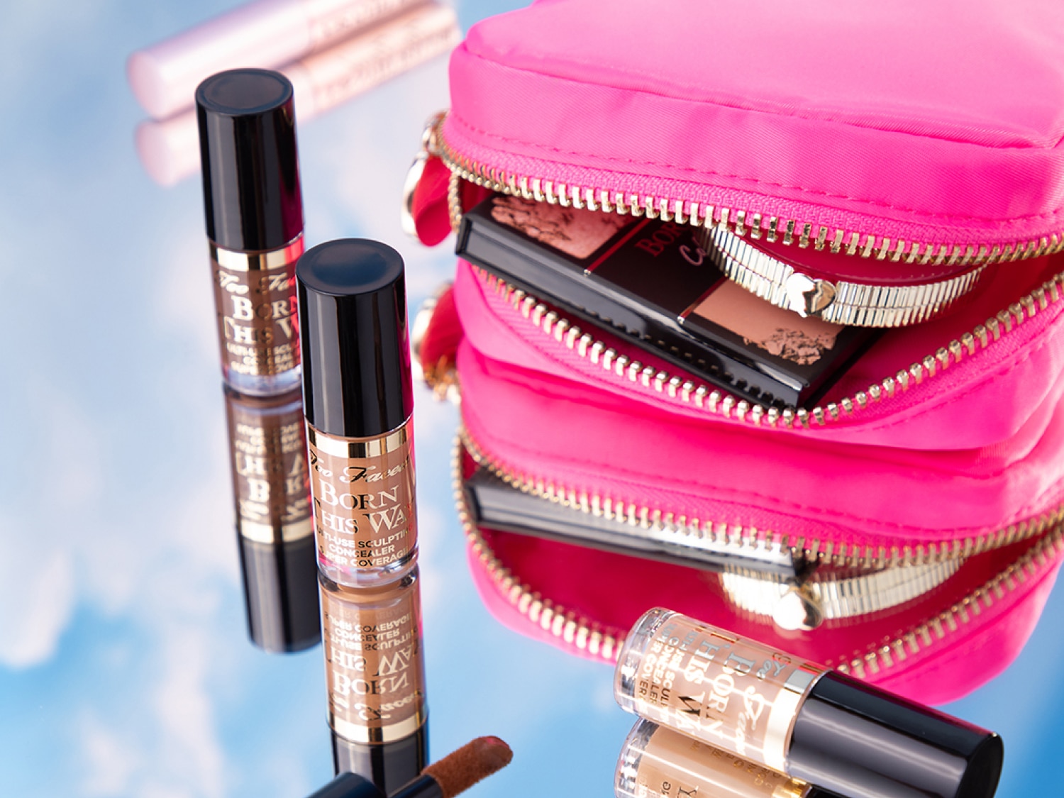 Auf einer reflektierenden Oberfläche liegen mehrere „Born This Way" Concealer in Travel-Size neben einer geöffneten, pinkfarbenen Kosmetiktasche, die Make-up-Produkte enthält.