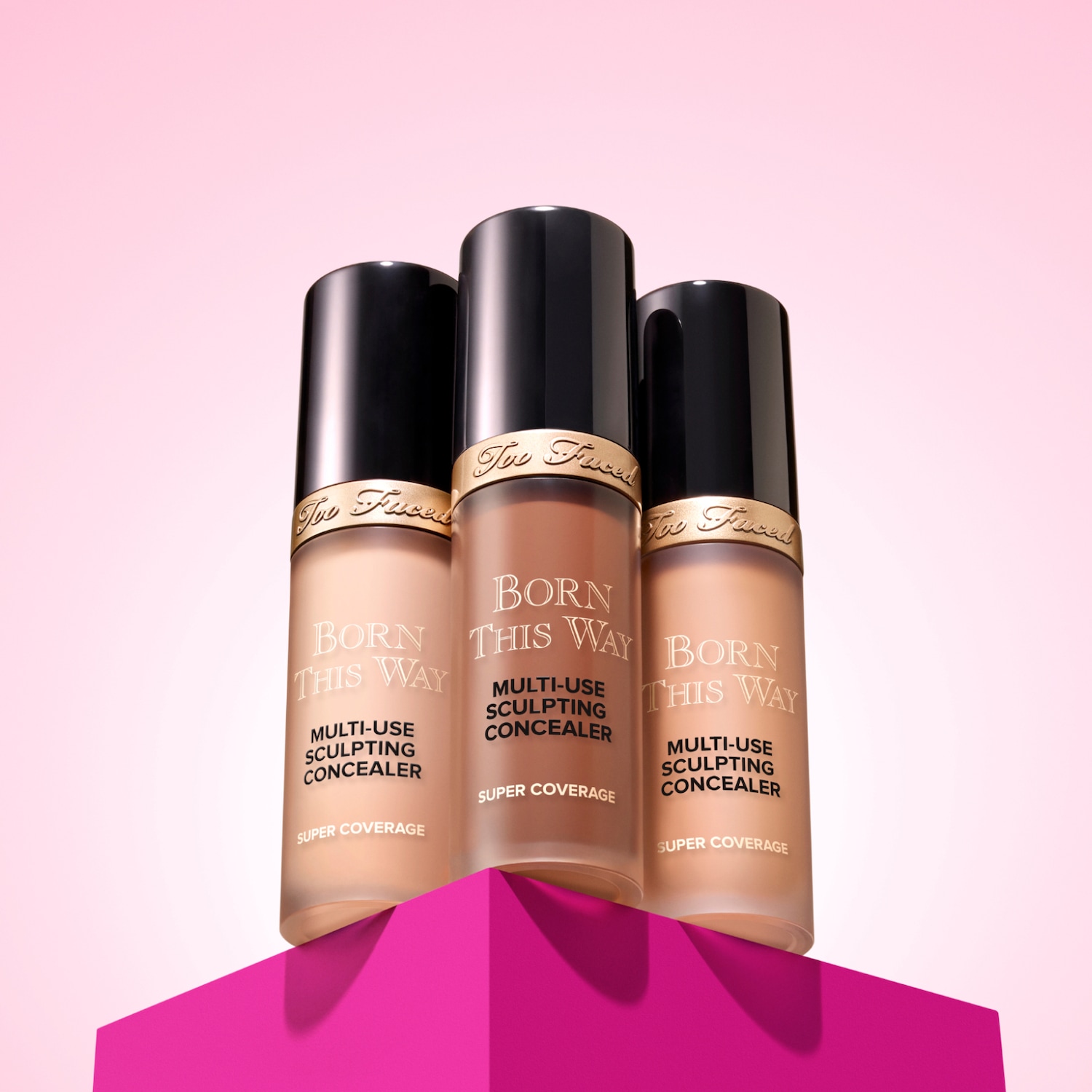 Drei Flaschen Born This Way Concealer von Too Faced mit schwarzem Deckel und verschiedenen Farbtönen stehen auf einem pinkfarbenen geometrischen Sockel.