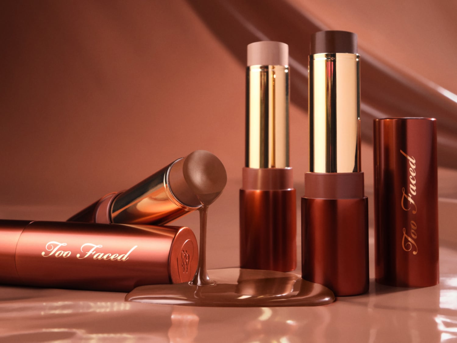 Mehrere Chocolate Soleil Melting Bronzing & Sculpting Sticks von Too Faced in verschiedenen Farbtönen stehen und liegen auf glänzendem, braunem Untergrund.