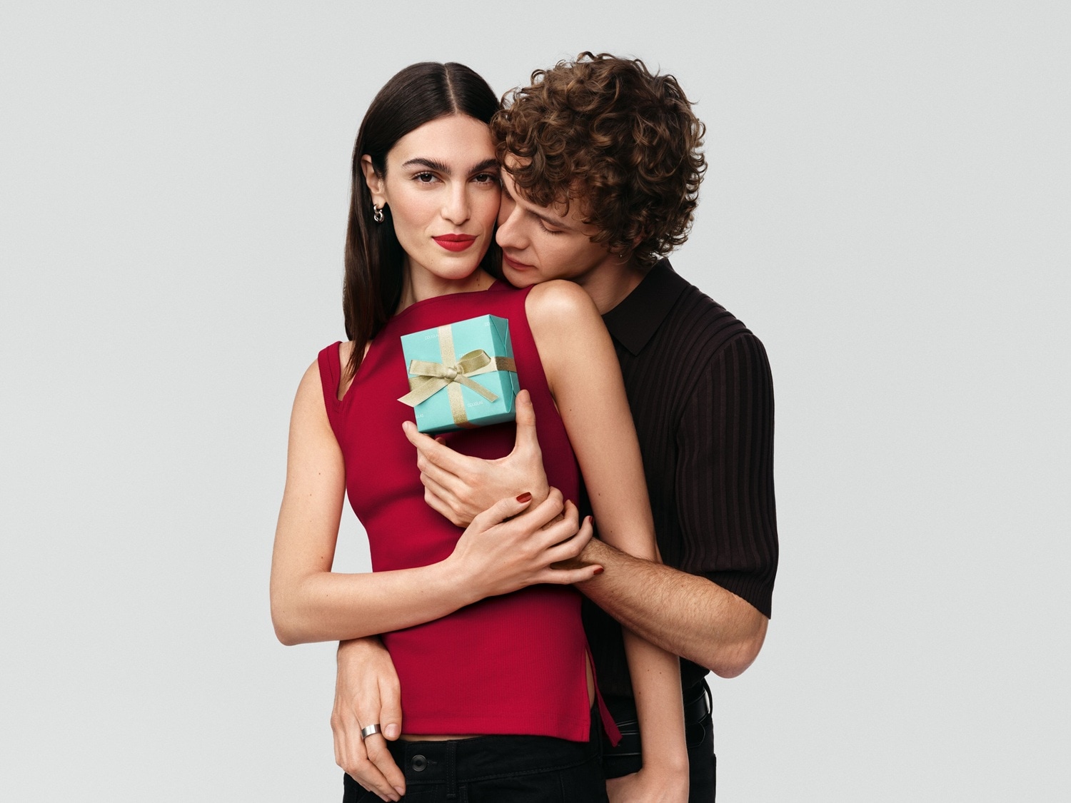 Ein Paar umarmt sich, die Frau hält eine kleine, hellblaue Geschenkbox mit einer goldenen Schleife, während der Mann sie von hinten umarmt.