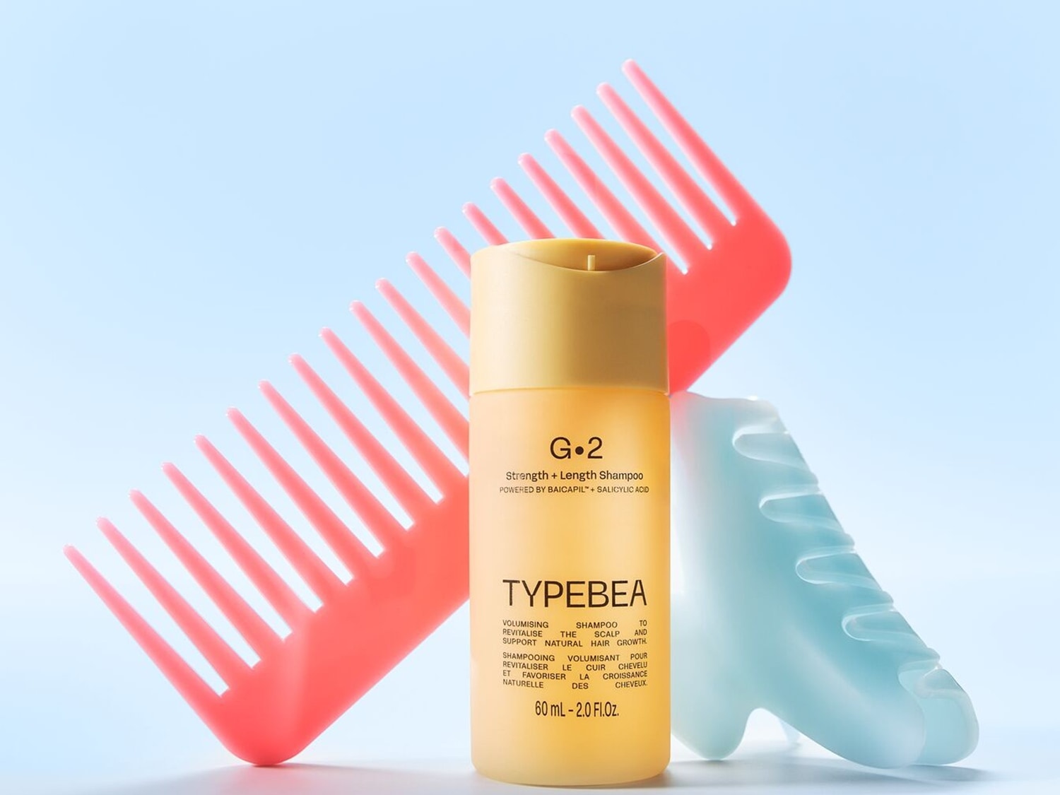G.2 Strength + Length Shampoo in een gele verpakking, met een roze kam en een blauwe haarborstel op de achtergrond, voor het revitaliseren van de hoofdhuid en het ondersteunen van de natuurlijke haargroei.