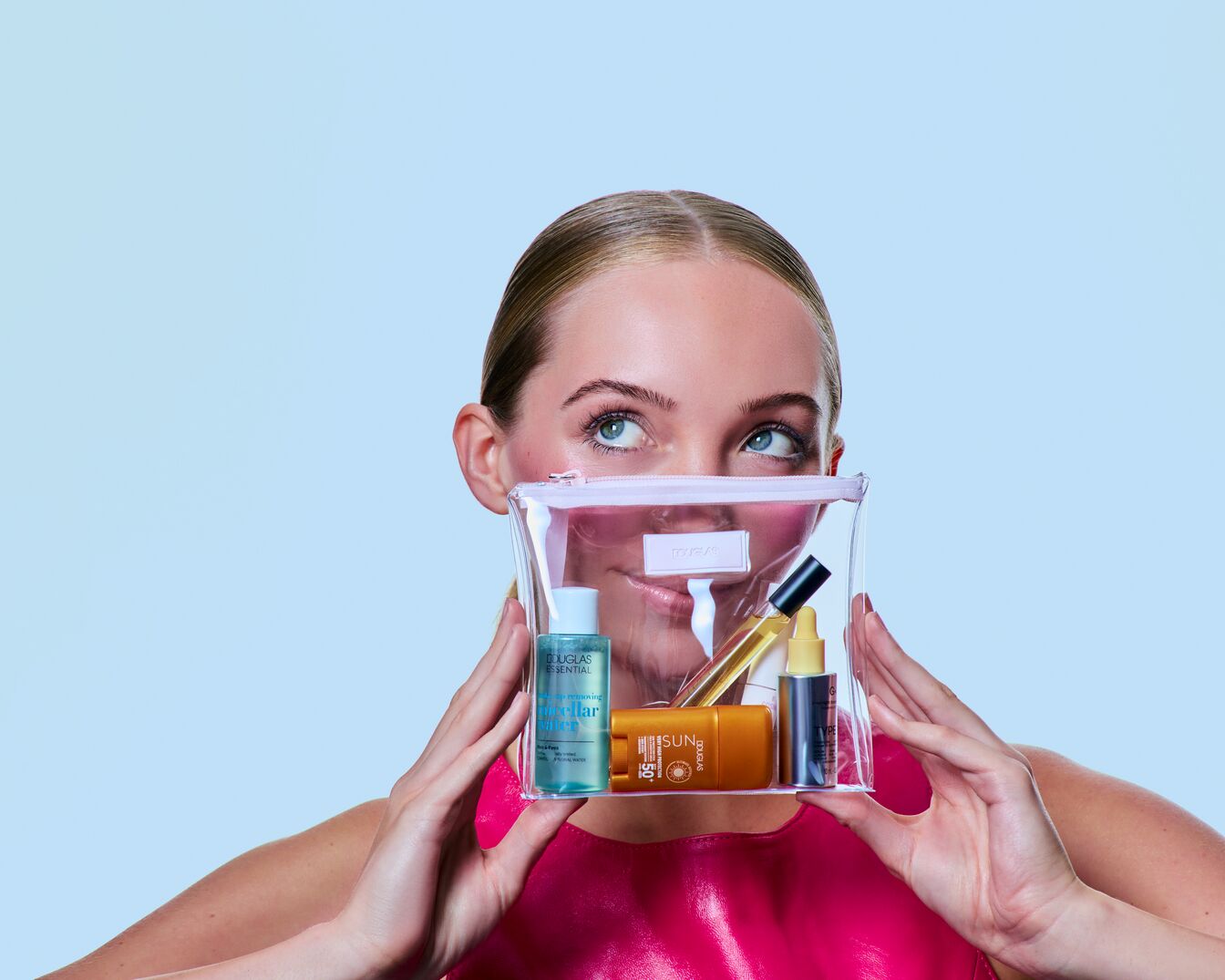 Een jonge vrouw houdt een transparant roze make-uptasje vast, gevuld met diverse Douglas schoonheidsproducten.