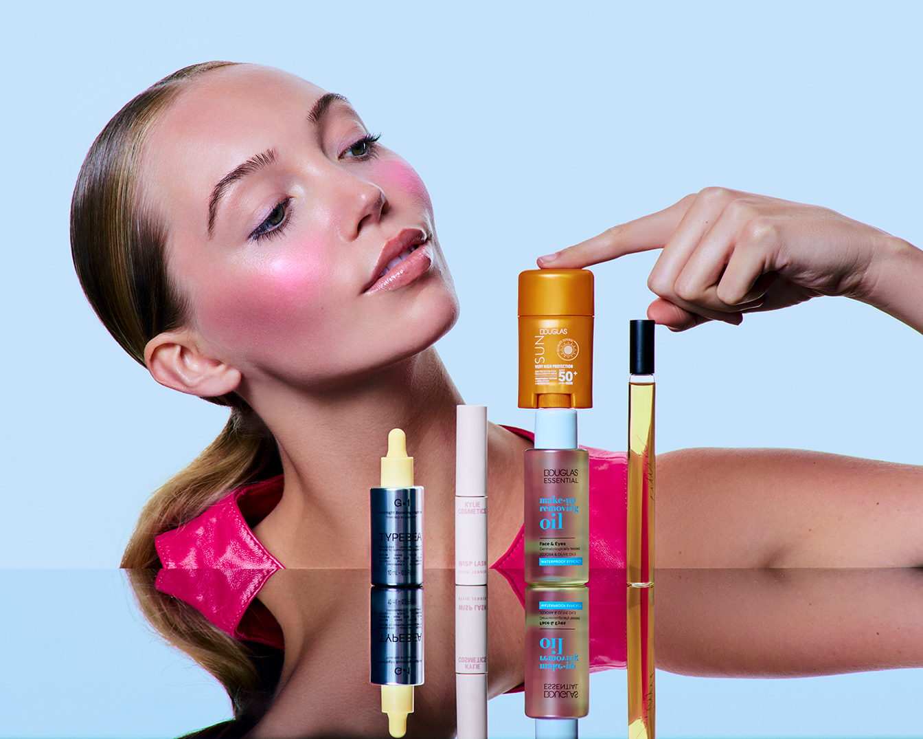 Na jasnoniebieskim tle, kobieta z różem na policzkach prezentuje serię produktów Douglas: olejek do demakijażu, tusz do rzęs Kylie Cosmetics, olejek i krem z filtrem SPF 50.