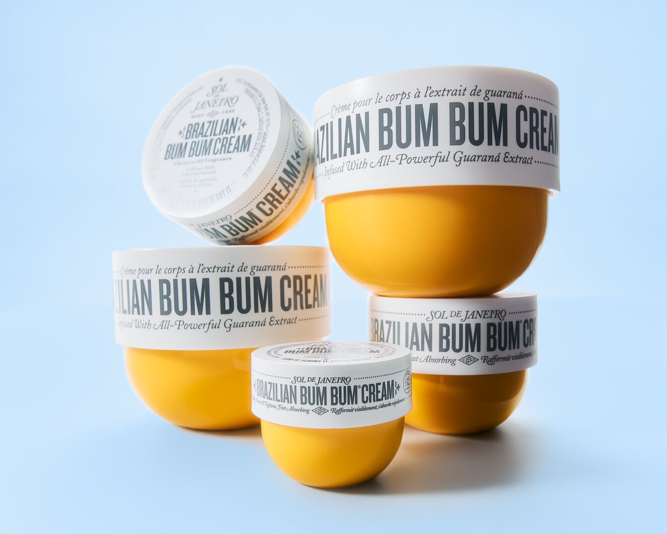 Stapel potten Sol de Janeiro Brazilian Bum Bum Cream, met witte deksels en gele containers, die de verstevigende en snel absorberende eigenschappen benadrukken.