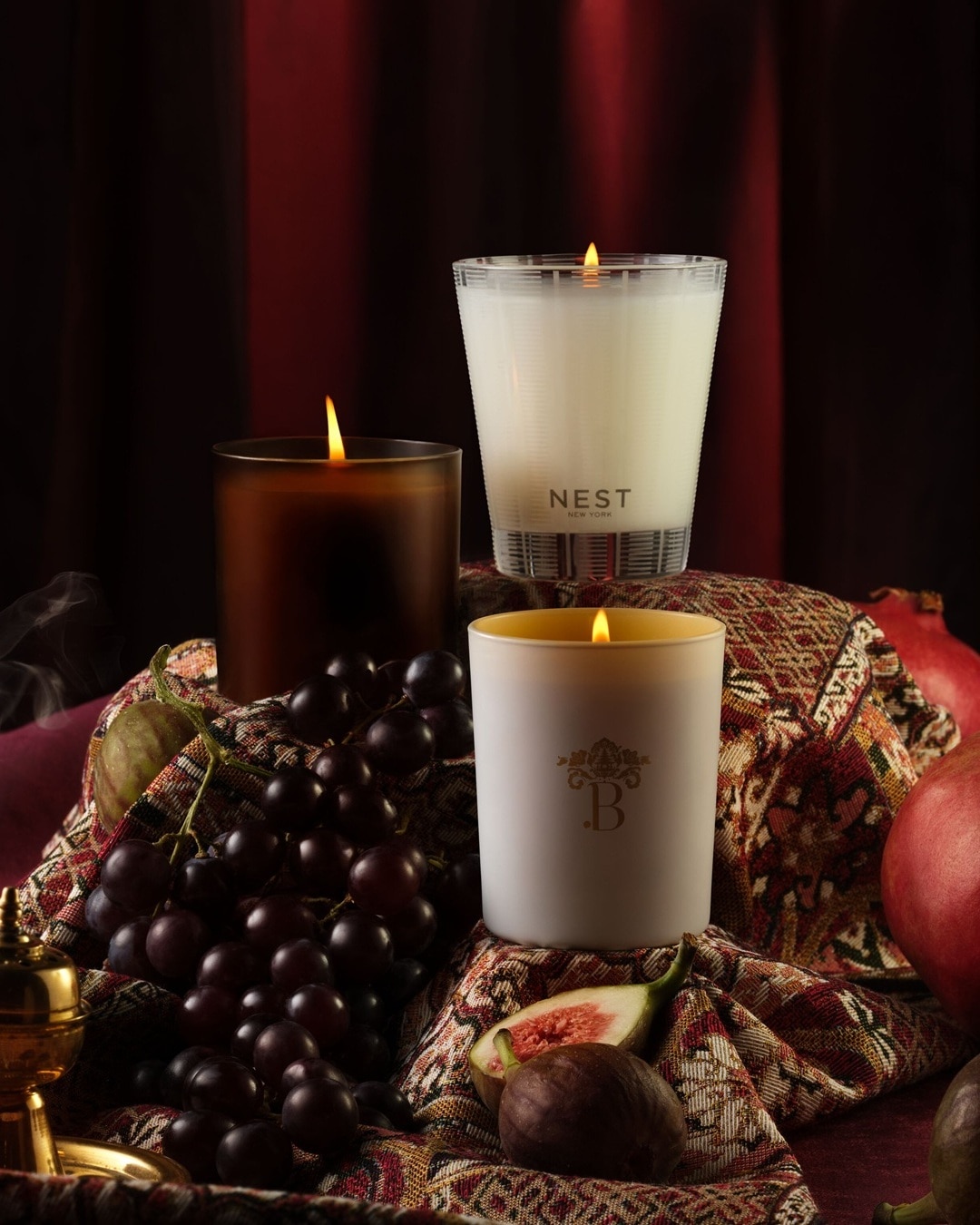 Drei Duftkerzen von Nest New York und einer weiteren Marke stehen auf einem Tuch mit Obst wie Trauben, Feigen und Granatäpfeln.