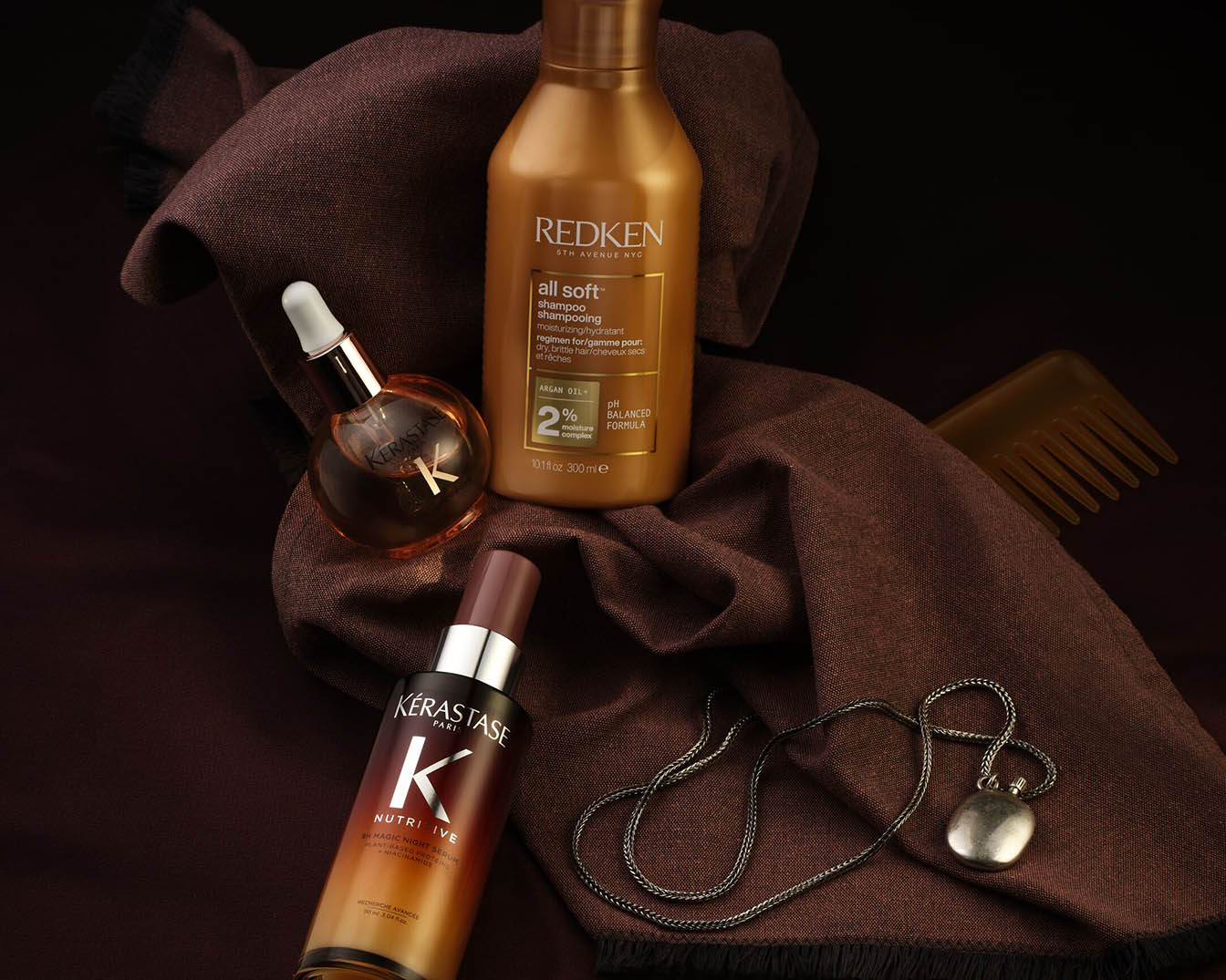 Ontdek de Redken All Soft shampoo in een gouden fles, samen met Kérastase Nutritive nachtserum en een haarolie, gepresenteerd op een bruine doek met een zilveren ketting en kam.
