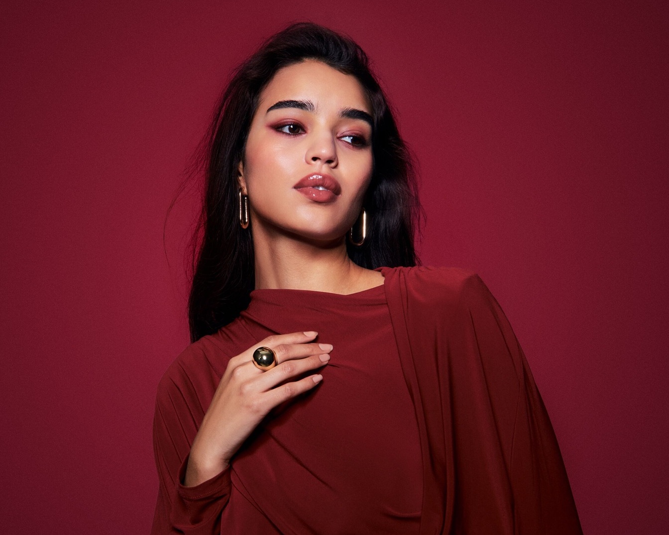 Een vrouw met donker haar, gouden oorbellen en een grote gouden ring, gekleed in een bordeauxrode jurk, kijkt weg tegen een bordeauxrode achtergrond.