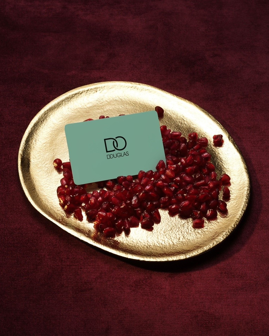 Auf einem goldenen Teller liegen rote Granatapfelkerne und eine hellblaue Douglas Geschenkkarte.