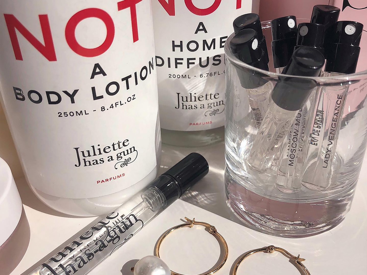 Entdecke Juliette has a Gun Parfums, inklusive Bodylotion, Raumduft und Duftproben in einem Glas, ergänzt durch goldene Ohrringe für einen Hauch von Luxus.