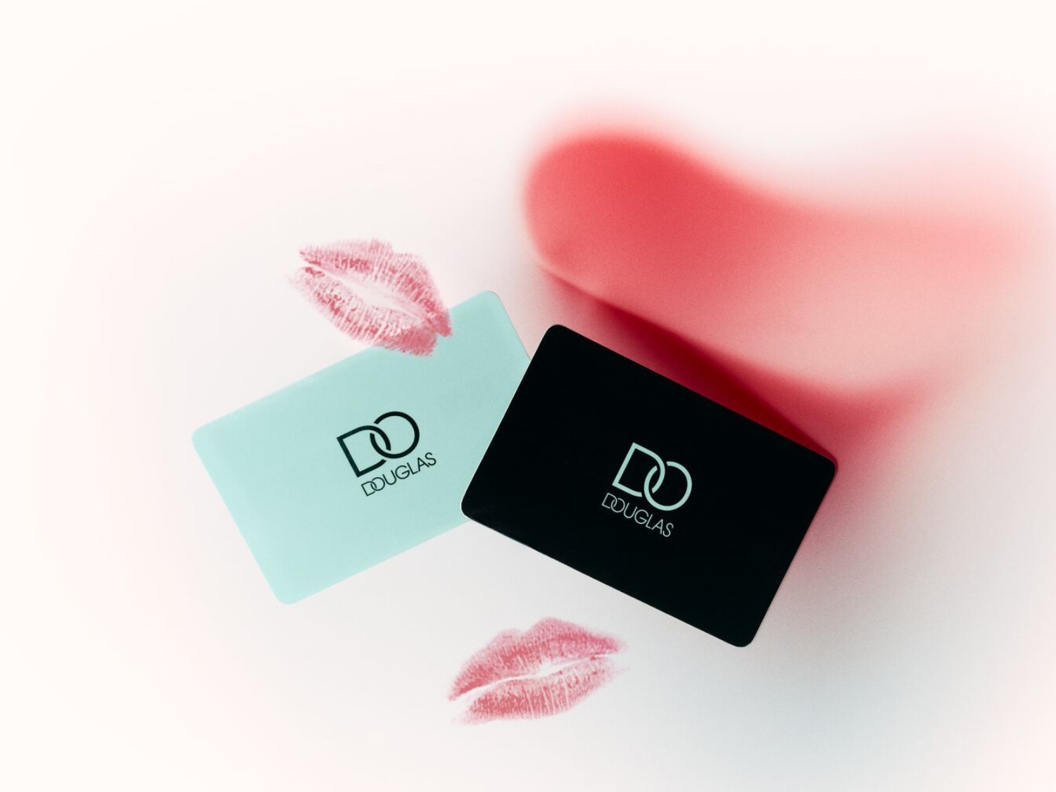 Dwie karty podarunkowe Douglas, jedna turkusowa, druga czarna, leżą na białym tle z odbitymi śladami ust.