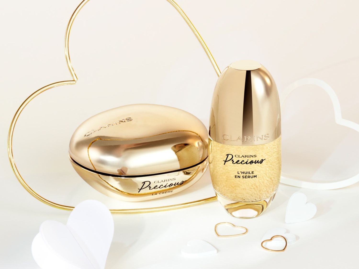 Découvrez le luxe avec Clarins Precious : La Crème dans son pot doré et L'Huile en Sérum dans son flacon élégant, présentés avec des décorations en forme de cœur.