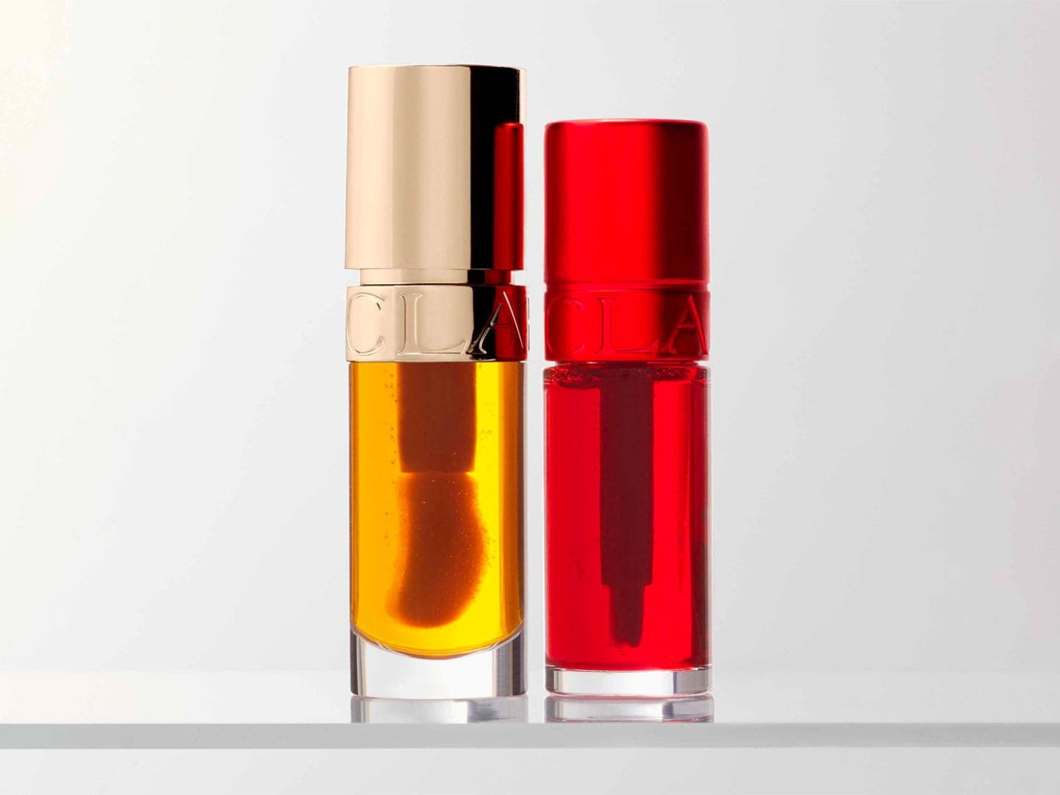 Voici deux huiles pour les lèvres Clarins, l'une dorée avec un bouchon beige et l'autre rouge vif, offrant une touche de couleur et de soin à vos lèvres.