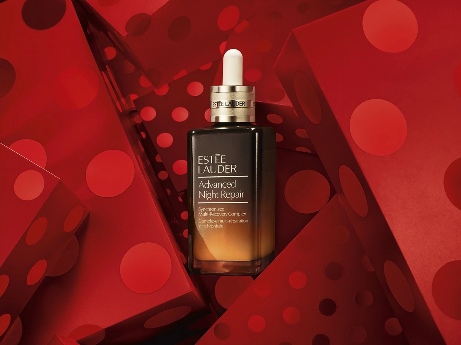 Découvrez le sérum Advanced Night Repair d'Estée Lauder, un complexe multi-réparation synchronisée dans un flacon dégradé noir et or, présenté sur un fond rouge à pois.
