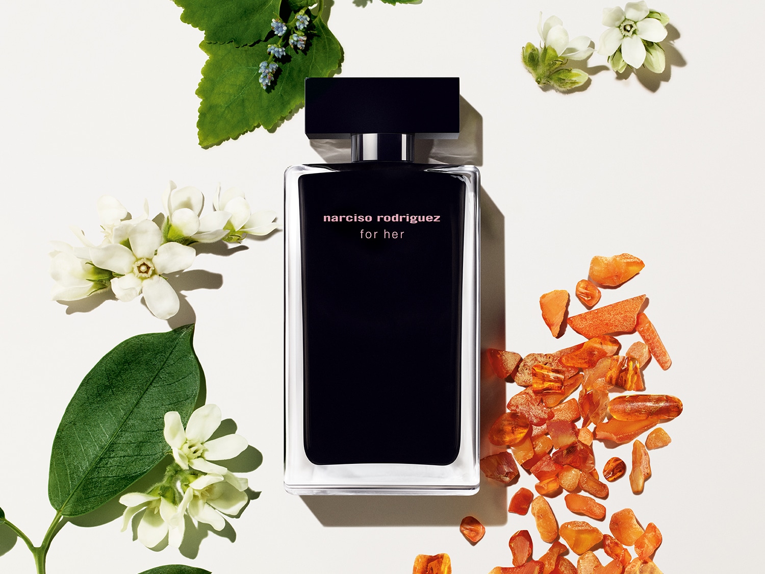 Schwarzer „Narciso Rodriguez for her“-Parfumflakon mit durchsichtigem Rand, umgeben von weißen Blüten, grünen Blättern und bernsteinfarbenen Steinen.