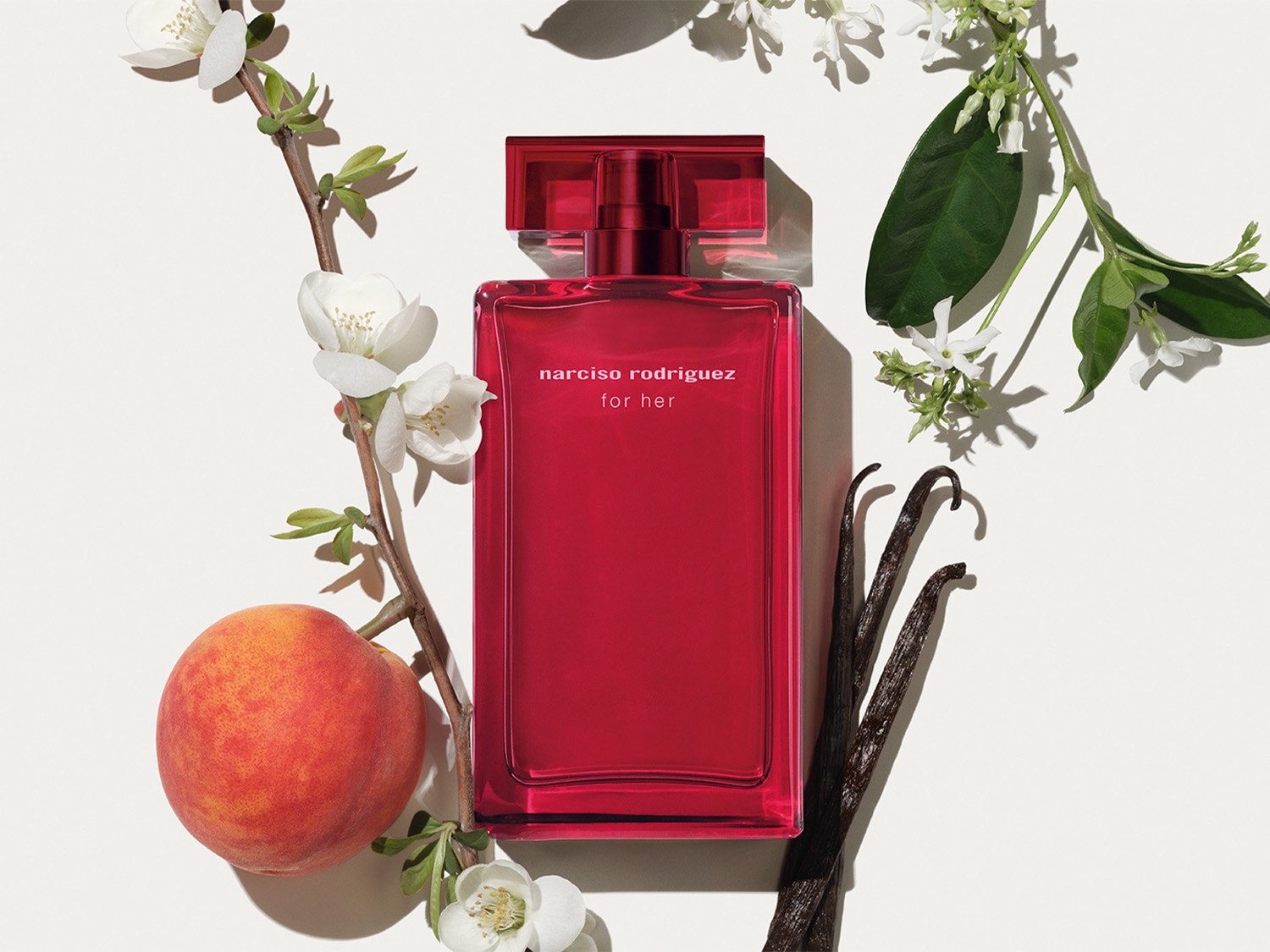 Ein roter Narciso Rodriguez For Her Parfümflakon steht neben einem Pfirsich, Vanilleschoten und weißen Blütenzweigen.