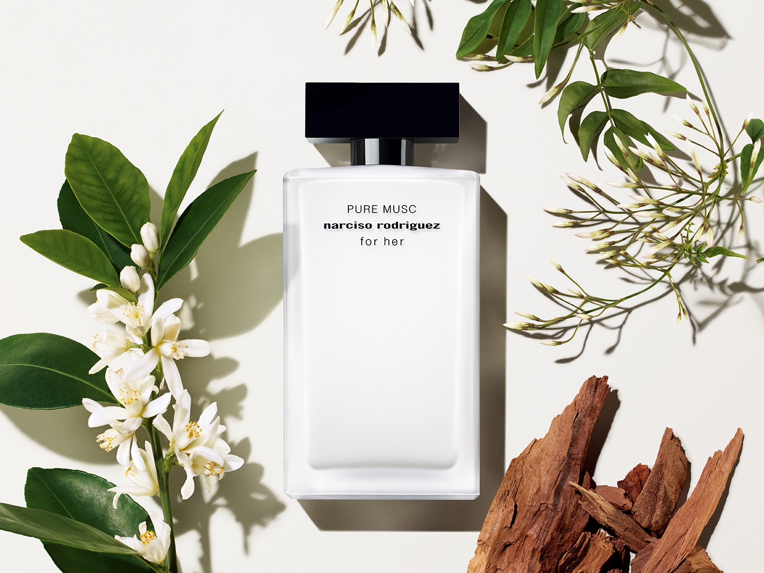 Ein weißer Flakon „for her Pure Musc“ von Narciso Rodriguez liegt inmitten von weißen Blüten, grünen Blättern und Holzstücken.