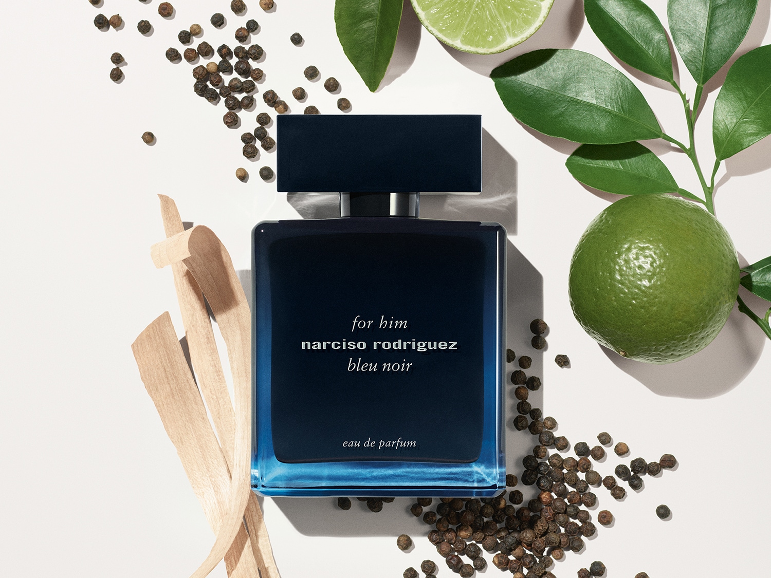 Eine Flasche Narciso Rodriguez Bleu Noir Eau de Parfum für Herren, umgeben von Limetten, Blättern, Holzspänen und Pfefferkörnern.