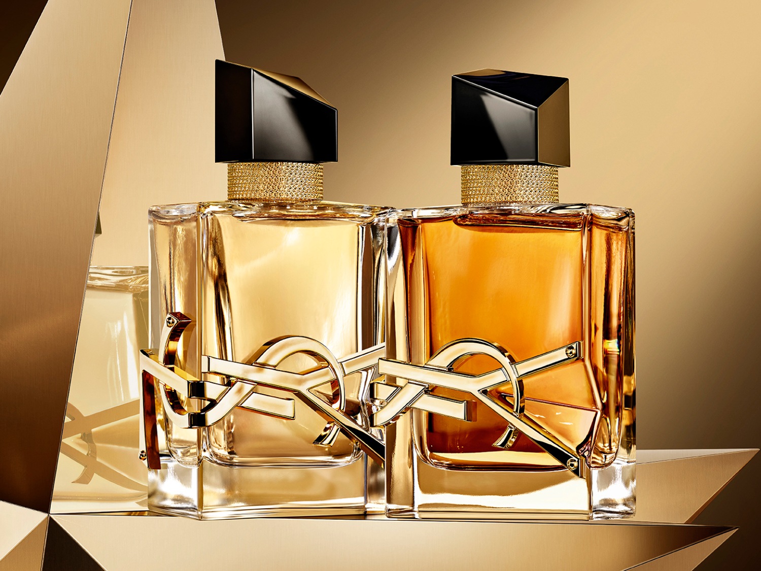 Twee vierkante parfumflessen van Yves Saint Laurent met gouden logo en zwarte dop, gevuld met vloeistoffen in lichte en donkere goudtinten.