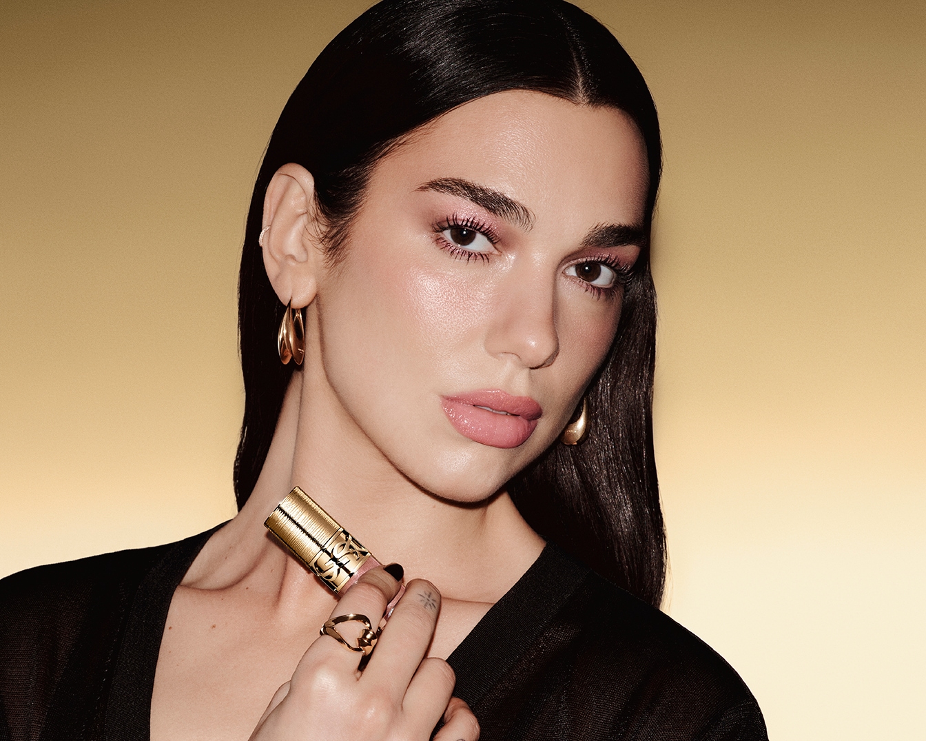Een vrouw met donker haar en gouden oorbellen houdt een gouden YSL-lippenstift vast, haar make-up is lichtroze.
