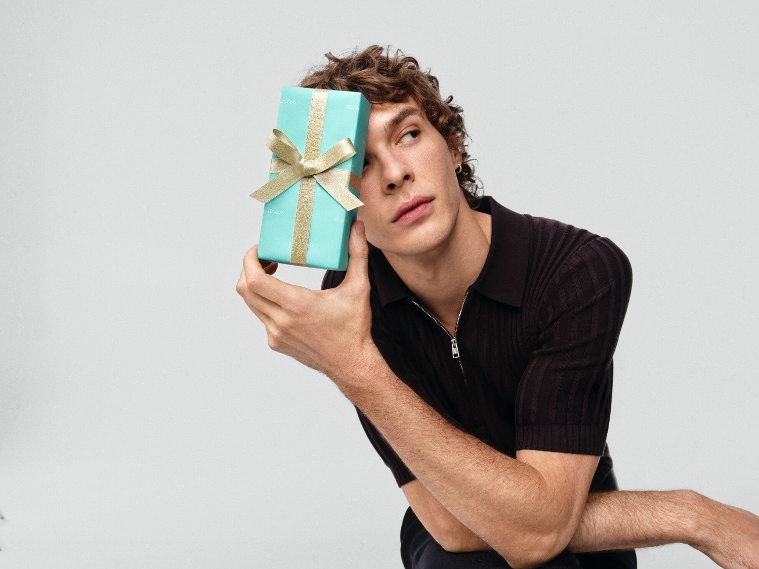 Mężczyzna z kręconymi włosami trzyma w dłoniach turkusowe pudełko prezentowe przewiązane złotą wstążką, spoglądając w bok.