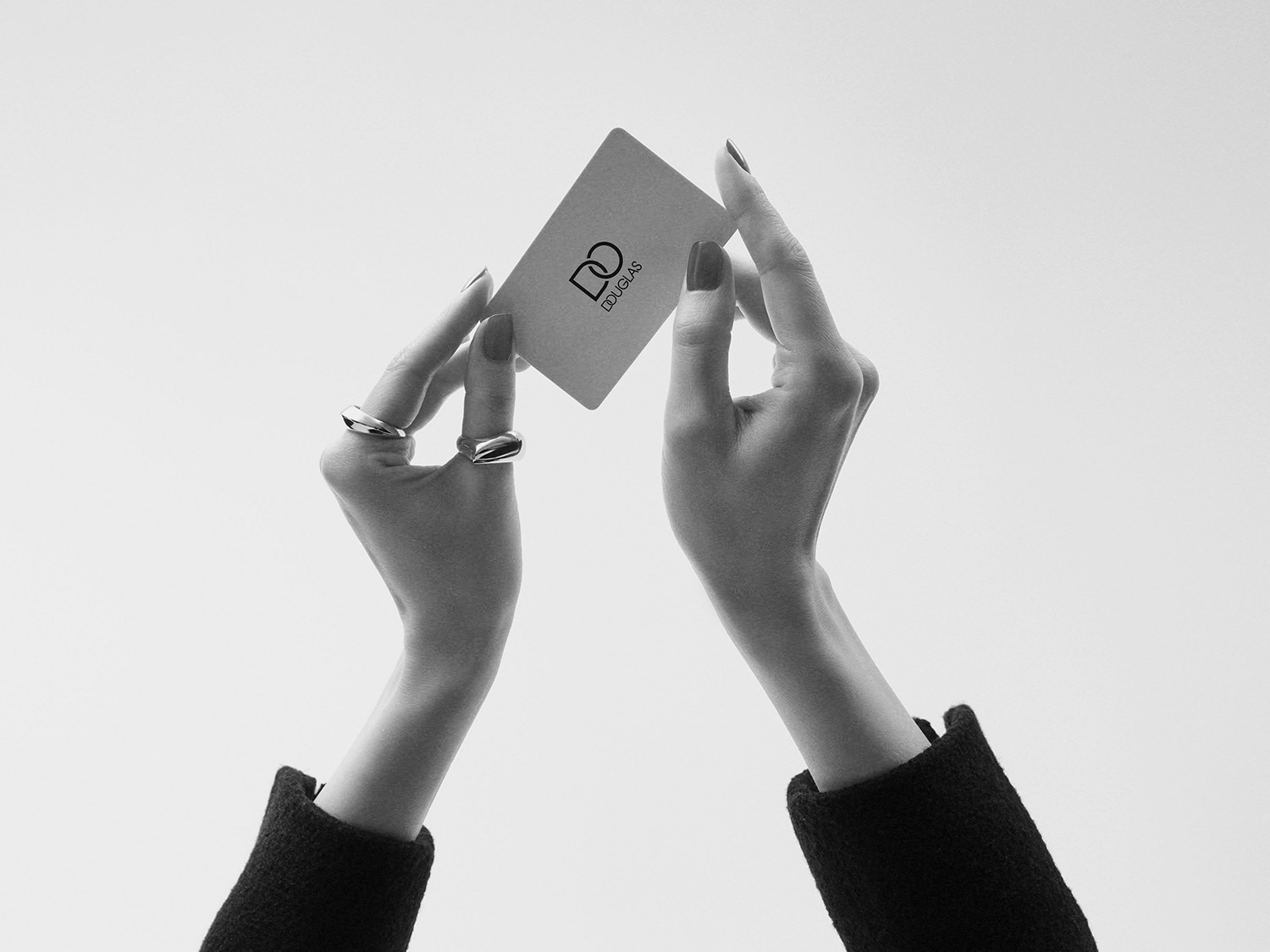 A woman's hands, adorned with rings and dark nail polish, hold up a Douglas gift card against a bright background.