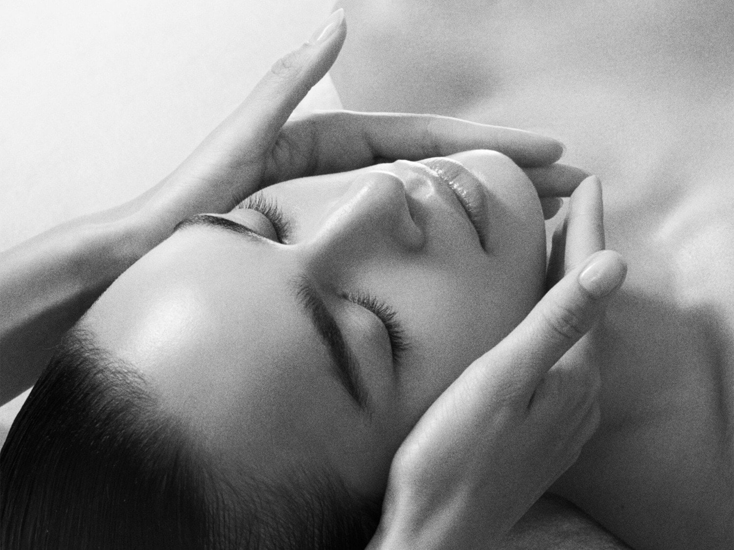 A serene black and white close-up captures a woman with closed eyes, her face gently framed by hands, suggesting a calming facial massage or skincare treatment.