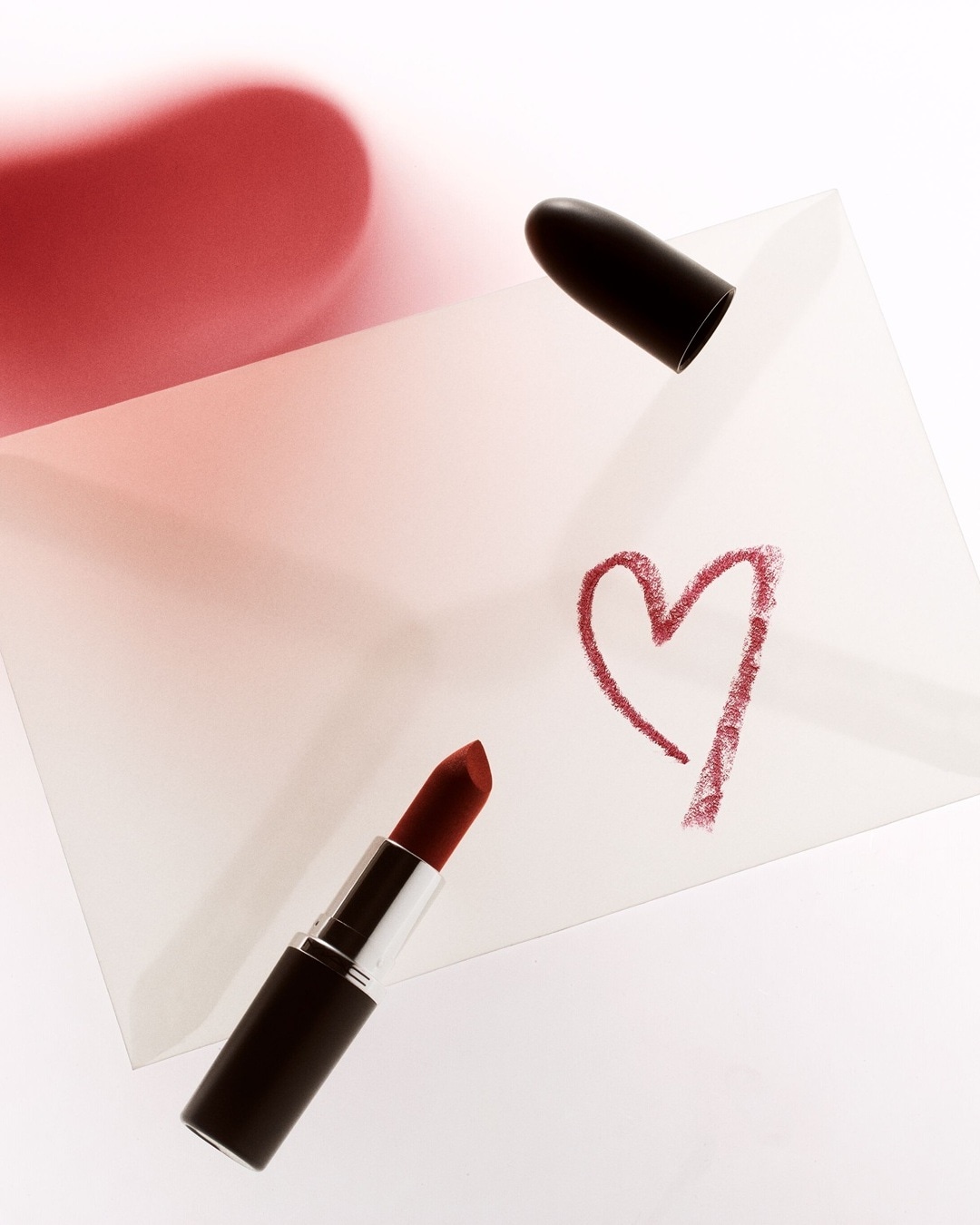Szminka w czarnym opakowaniu leży obok narysowanego serca na białym tle, tworząc romantyczny akcent.