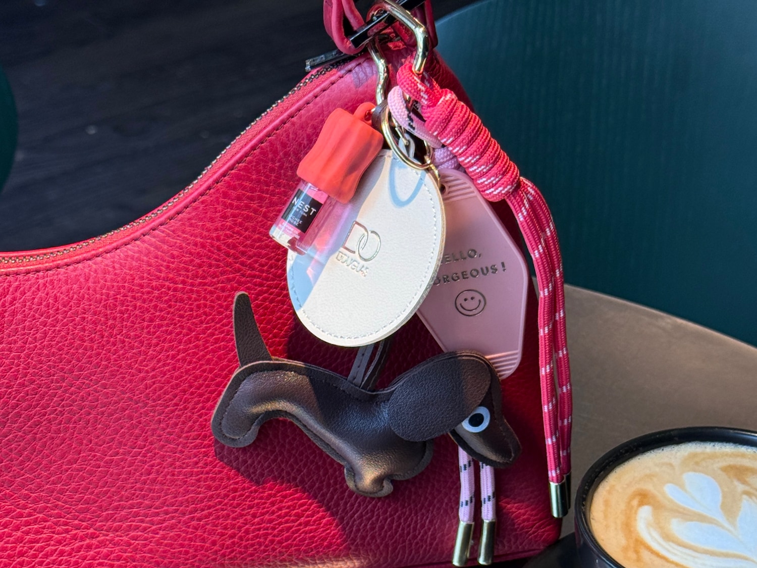 Nahaufnahme einer roten Handtasche mit Anhängern, darunter ein Dackel, ein Nest-Parfüm und ein Douglas-Logo.