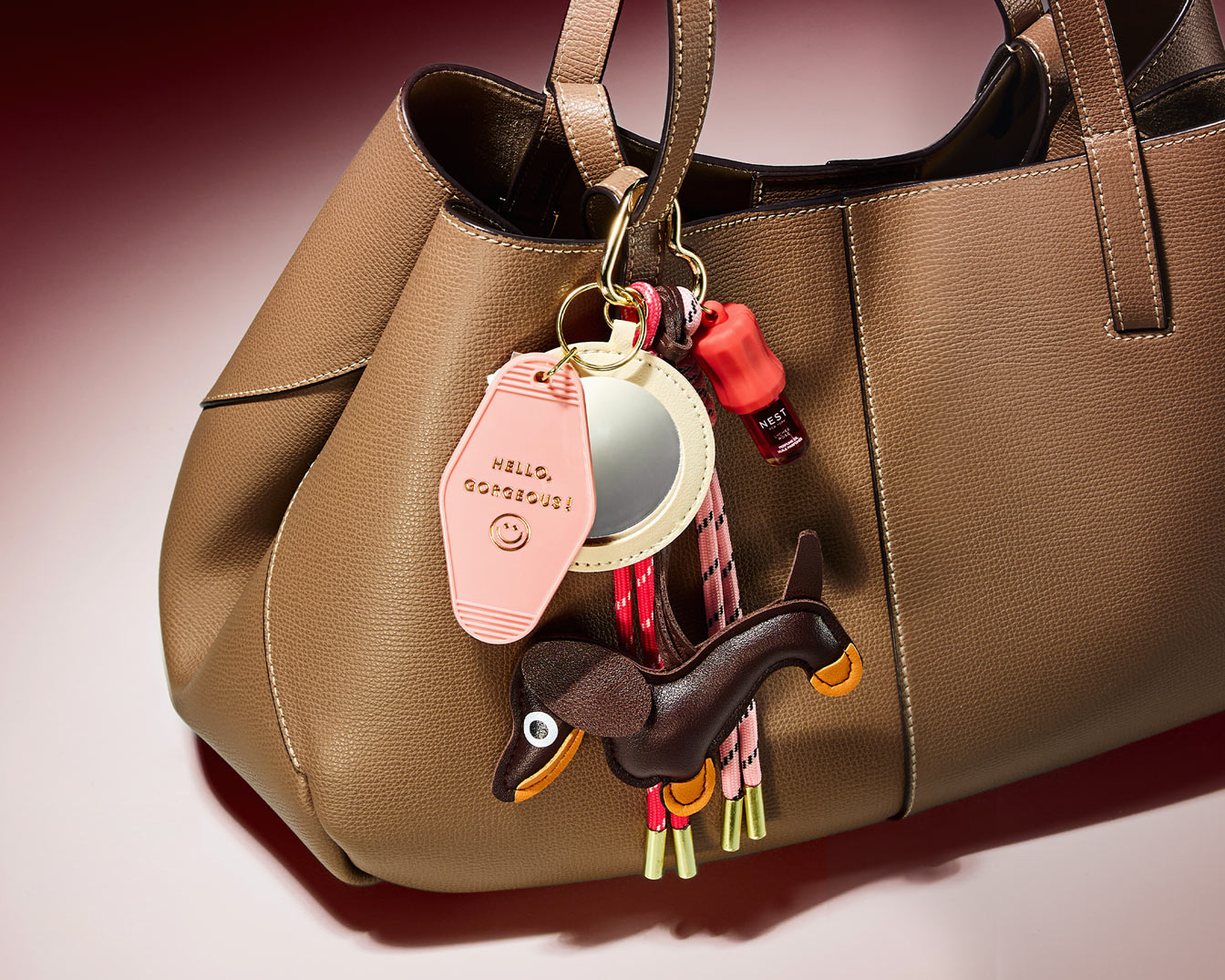 Nahaufnahme einer braunen Handtasche mit Anhängern, darunter ein rosa Schlüsselanhänger mit der Aufschrift "Hello, Gorgeous!", ein Spiegel, ein Nagellack von Nest und ein Dackel.