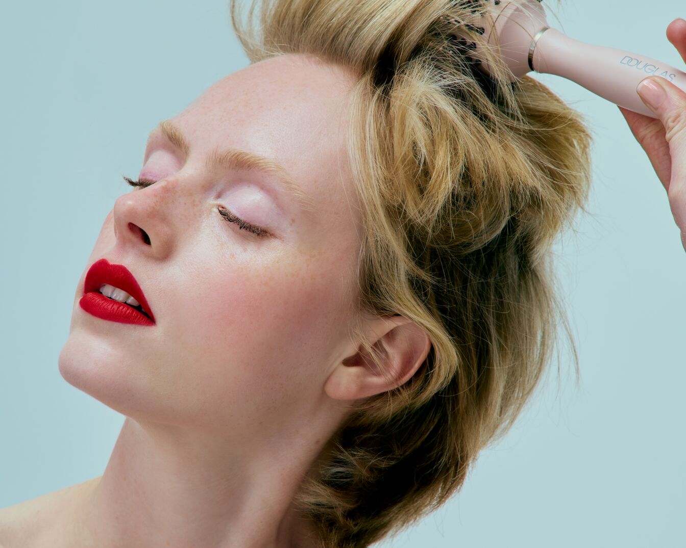 Eine blonde Frau mit geschlossenen Augen und roten Lippen wird mit einer rosafarbenen Douglas Rundbürste frisiert.