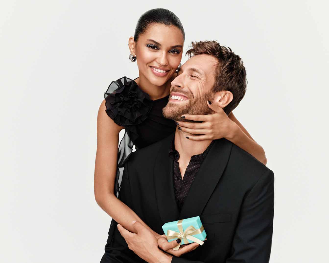 Uśmiechnięta kobieta w czarnej sukience obejmuje mężczyznę w garniturze, który trzyma małe, błękitne pudełko z kokardką.