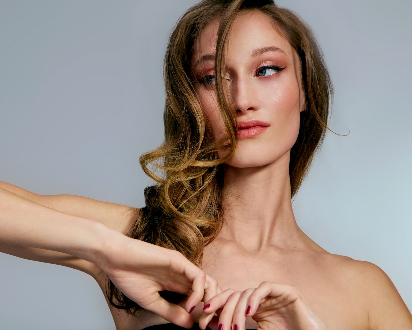 Portret kobiety z długimi, kręconymi włosami w odcieniach blondu, z delikatnym makijażem oczu i ust, pozującej na szarym tle.