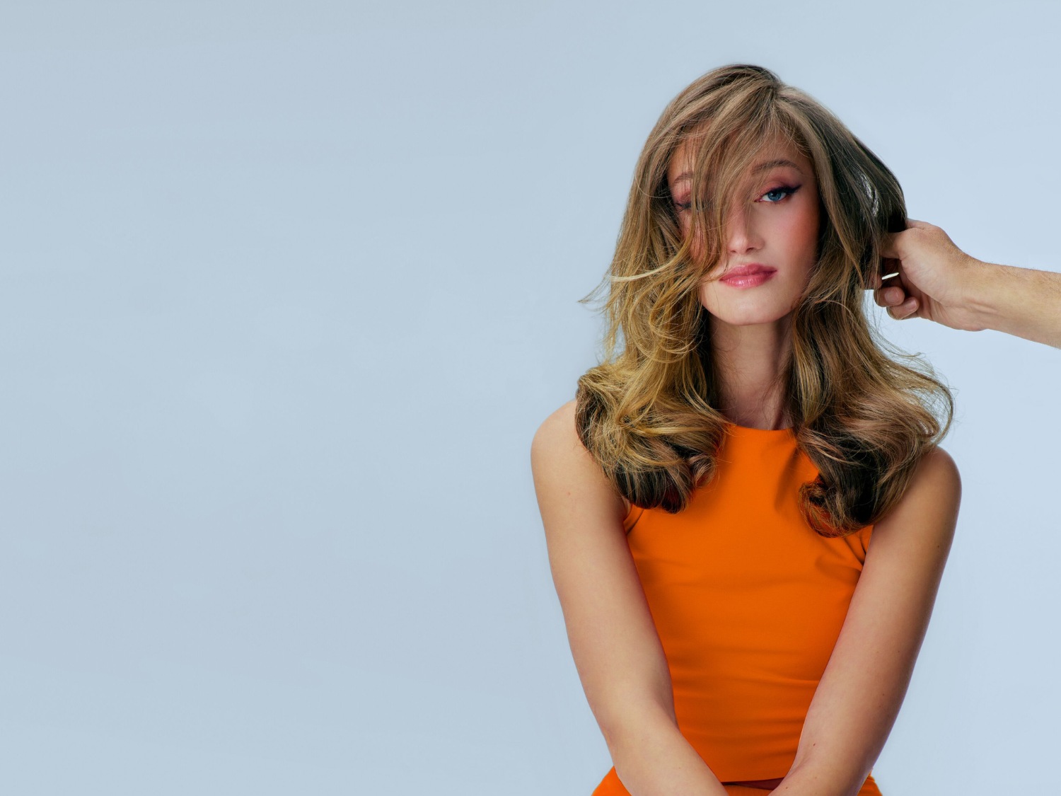 Na jasnym tle kobieta z długimi, falowanymi włosami w odcieniach blondu i brązu, ubrana w pomarańczową bluzkę, pozuje z ręką stylisty układającą jej włosy.