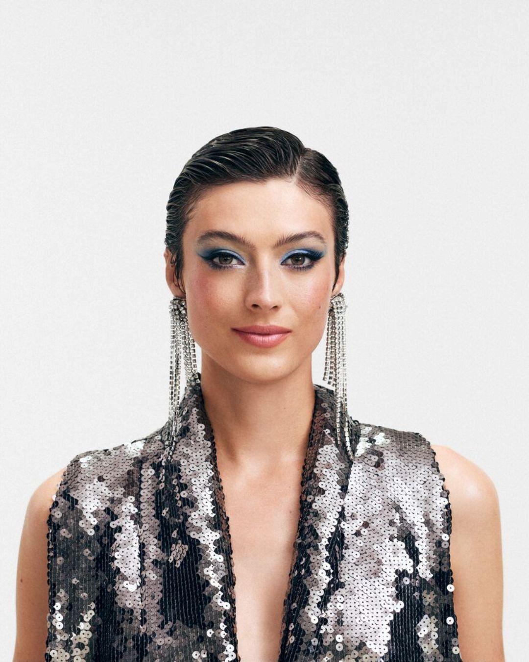 Primo piano di una modella con ombretto blu brillante, orecchini pendenti argentati e un abito senza maniche con paillettes bianche e nere.