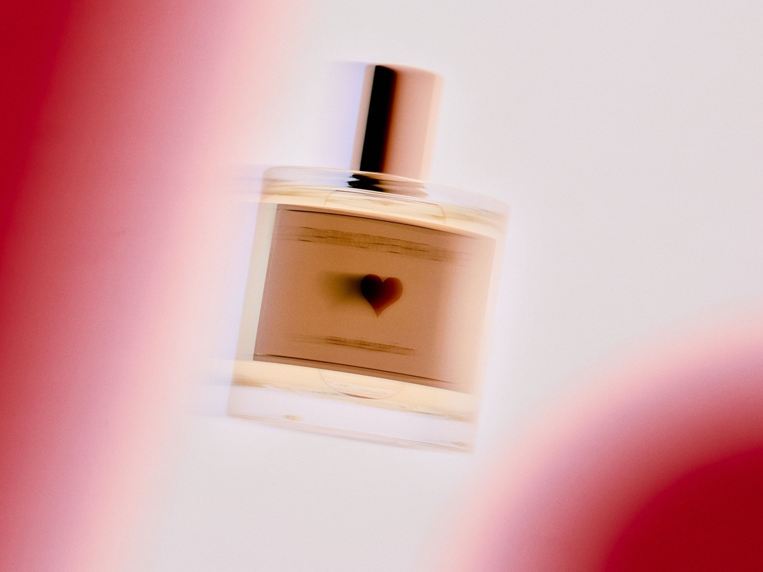 Przezroczysty flakon perfum z beżową etykietą ozdobioną czarnym sercem i złotą zatyczką, otoczony rozmytymi czerwonymi kształtami.