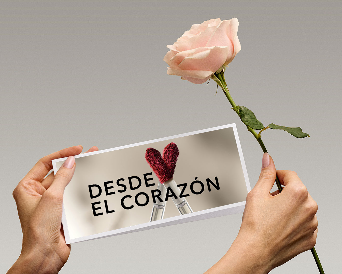Una mano sostiene una foto con el texto 'Desde el Corazón' y un corazón rojo difuso, mientras que la otra mano sostiene una rosa rosa pálido, creando un ambiente romántico y delicado.